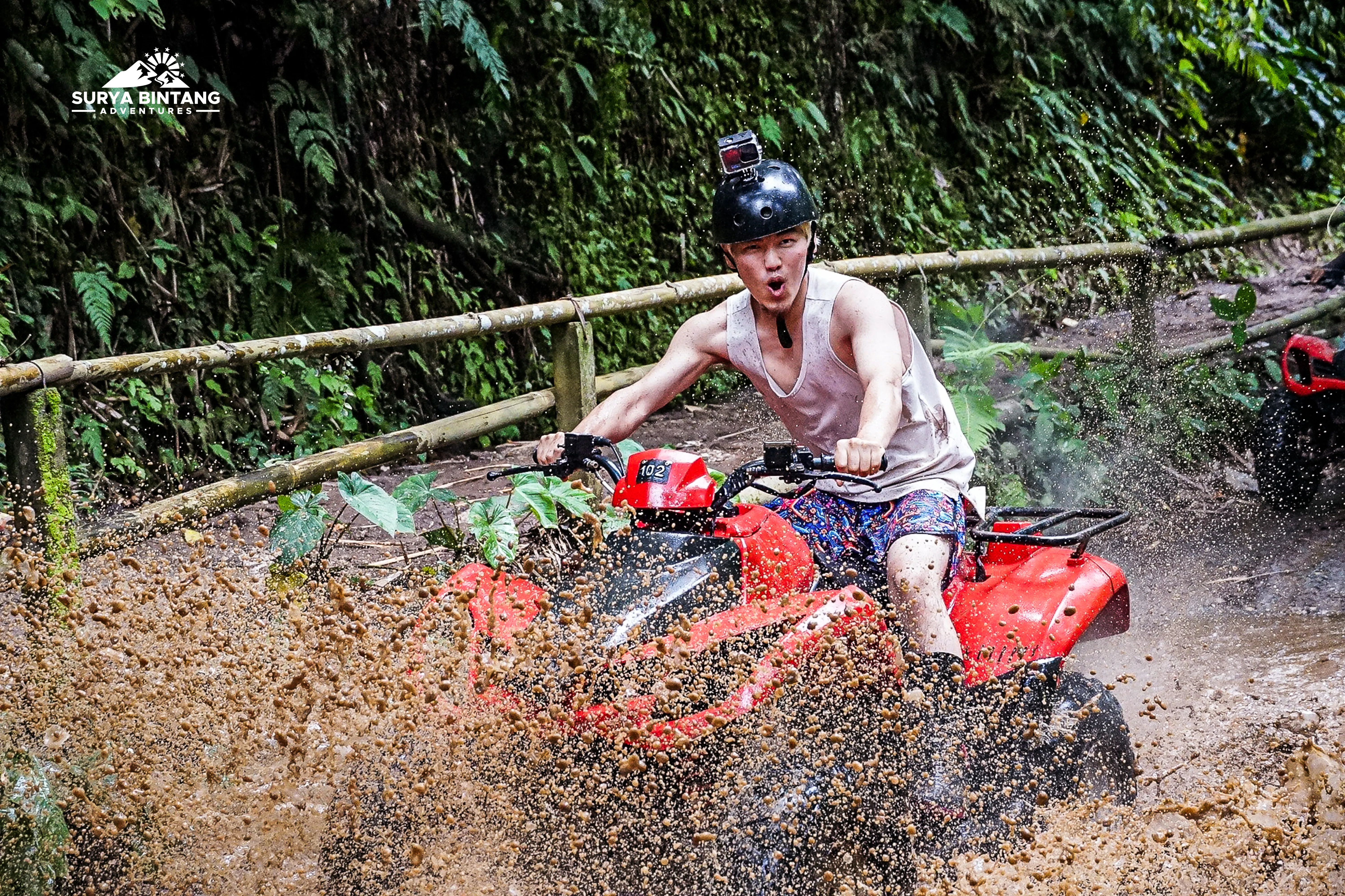 Young man riding a red ATV through a muddy trail in a lush green forest