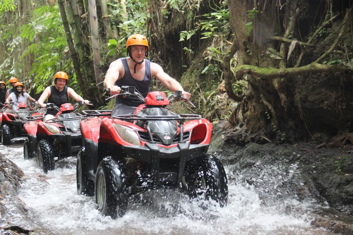 Group of people riding red ATVs through a shallow forest stream surrounded by lush green vegetation